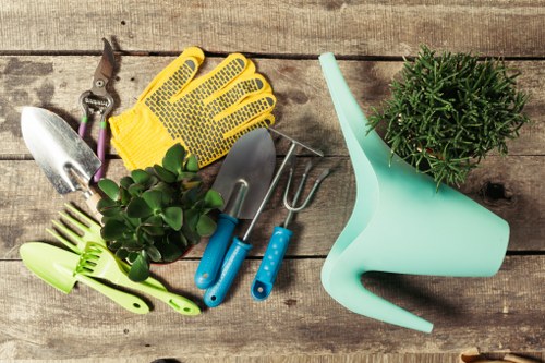 The image displays an assortment of gardening tools and accessories arranged on a weathered wooden surface. Included are a pair of yellow rubber gloves with textured palms, a small hand trowel with a metallic blade and wooden handle, a pair of pruning shears, a green hand rake, and two blue-handled cultivator tools. Additionally, there is a light blue plastic watering can positioned on its side, with a small potted shrub featuring green, needle-like foliage placed nearby. The scene suggests a gardening setup suitable for maintaining a garden or lawn, with tools ready for planting, weeding, watering, and general outdoor maintenance. The wooden surface provides a rustic backdrop, indicative of outdoor storage or preparation for gardening work, relevant to services offered by Lawn Mowing Euston and local gardening professionals in the Euston area, within postal code NW1 or nearby. The lighting appears natural, suggesting an outdoor environment with soft, diffuse daylight, contributing to a calm and professional presentation of gardening essentials.