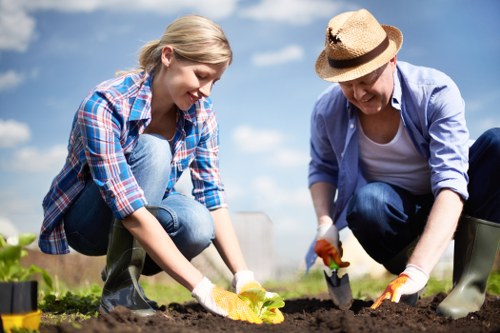 A young woman and an older woman are working together in a well-maintained garden, planting or tending to the soil using small gardening tools. The garden features a lush, green lawn bordered by a flower bed with dark, rich soil, and is set in an outdoor environment with a bright blue sky and a few clouds visible overhead. The women are dressed in casual gardening attire, including gloves and rubber boots, suggesting they are engaged in outdoor maintenance or planting activities. The garden layout includes a defined pathway or border separating the lawn from the flower bed, and in the background, there are some small plants or shrubs. The sunlight illuminates the scene, highlighting the natural tones of the earth, plants, and their clothing, emphasizing a productive gardening session under clear weather. This image aligns with professional gardening services such as those offered by Lawn Mowing Euston, specialising in lawn and garden maintenance around the Euston area.